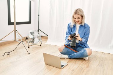 Fotoğrafçılık stüdyosunda çalışan güzel sarışın kadın yerde oturmuş bilgisayardaki fotoğrafları kontrol ediyor.