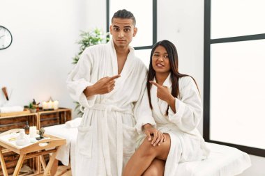 Young latin couple wearing towel standing at beauty center pointing aside worried and nervous with forefinger, concerned and surprised expression 