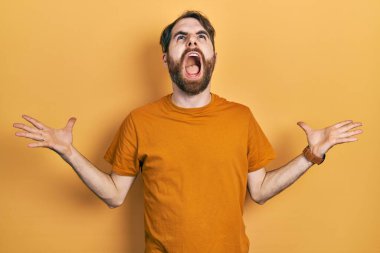 Sakallı, sıradan sarı tişörtlü, çılgın ve sinirli bir beyaz adam agresif bir ifadeyle bağırıyor ve kollarını kaldırıyor. hüsran kavramı. 