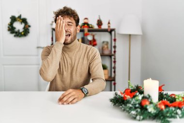 Masada sakallı genç yakışıklı bir adam Noel süslemesi ile oturuyor bir gözünü eliyle kapatıyor, yüzünde kendine güvenen bir gülümseme ve sürpriz bir duygu var.. 