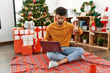 Arap genç adam dizüstü bilgisayar kullanıyor Noel ağacının yanında oturmuş parmaklarını göstererek reklamını yapıyor, şaşırmış yüzünü ve ağzını açıyor. 