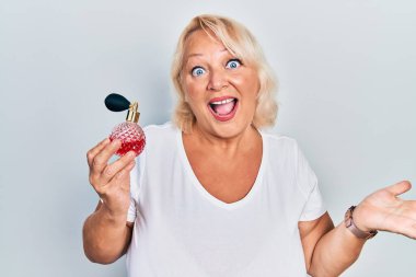 Orta yaşlı sarışın kadın parfüm tutuyor başarıyı kutluyor mutlu bir gülümseme ve kazanan yüz ifadesi kaldırılmış bir el ile 