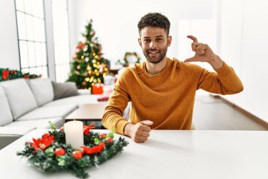 Arap genç adam Noel ağacının yanında oturmuş gülümsüyor ve el hareketleriyle küçük boy işareti yapıyor parmaklarıyla ve kamerasıyla kendine güveniyor. ölçüm kavramı. 