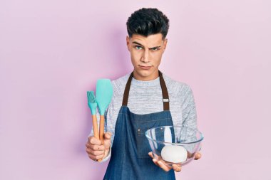 Genç İspanyol adam ekmek hamuru ve yemek aletleri tutuyor şüpheci ve gergin, sorun yüzünden surat asıyor. negatif kişi. 