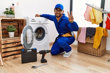 Çamaşır makinesinde çalışan genç Hintli teknisyen kameraya gülümseyen gülen yüzle parmaklarıyla zafer işareti yapıyor. İki numara.. 