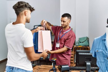 Genç İspanyol esnaf gülümseyerek giyim mağazasındaki müşteriye alışveriş çantası veriyor..