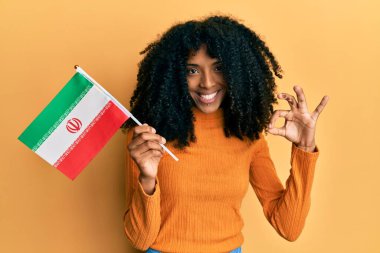Afro saçlı, Kürdistan bayrağı tutan Afro-Amerikalı kadın parmaklarıyla tamam işareti yapıyor, gülümseyerek mükemmel bir sembol oluşturuyor. 