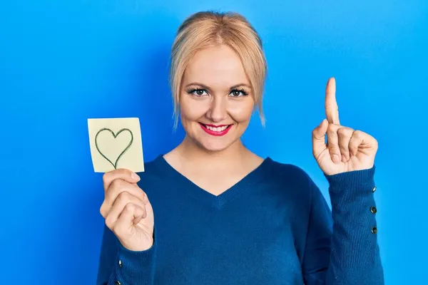 Mujer Rubia Joven Sosteniendo Recordatorio Del Corazón Sonriendo Con Una — Foto de Stock