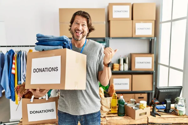 Gönüllü olarak bağış kutusunu tutan yakışıklı, orta yaşlı bir adam gülümseyen bir yüz ifadesiyle ayakta duruyor ve başparmağını kaldırarak yüzünü işaret ediyor.. 