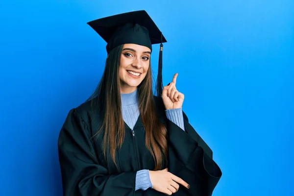 美しいブルネット若い女性を身に着けています卒業キャップと式のローブ顔に大きな笑顔で カメラを見て側に手と指で指して — ストック写真