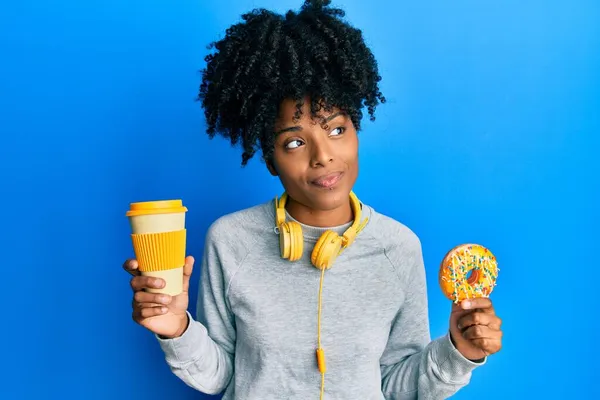 黑头发的非洲裔美国女人吃着甜甜圈 喝咖啡时面带微笑地看着旁边 目不转睛地想着 — 图库照片