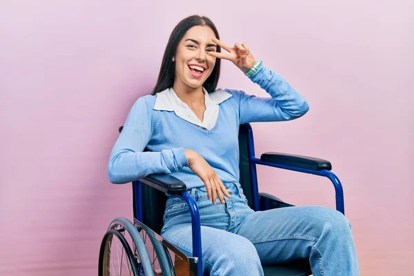 青い目をした美しい女性が車椅子に座り 顔の上に指で平和の象徴をして 陽気に勝利を示す笑顔 — ストック写真