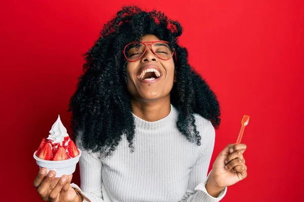 African american woman with afro hair eating strawberry ice cream smiling and laughing hard out loud because funny crazy joke.
