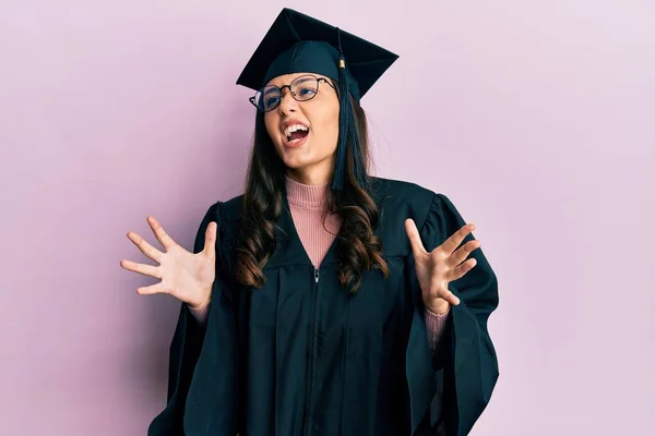 卒業キャップと式のローブを身に着けている若いヒスパニック系の女性は狂気と怒りの叫びと積極的な表現と腕を上げて叫んでいた フラストレーションコンセプト — ストック写真