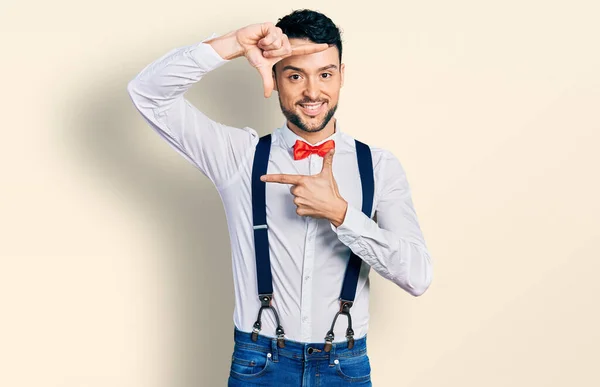 Hispanic man with beard wearing hipster look with bow tie and suspenders smiling making frame with hands and fingers with happy face. creativity and photography concept.