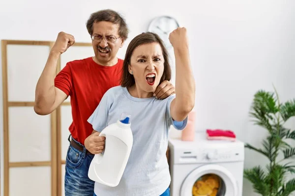 Międzyrasowa Para Średnim Wieku Robi Pranie Trzymając Butelkę Detergentu Rozdrażniona — Zdjęcie stockowe