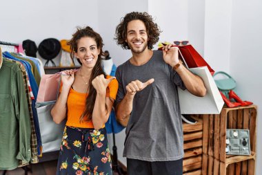 Perakende dükkanında alışveriş torbaları tutan genç İspanyol çift arka tarafı işaret ediyor. El ve baş parmakları havada, kendinden emin gülümsüyor. 