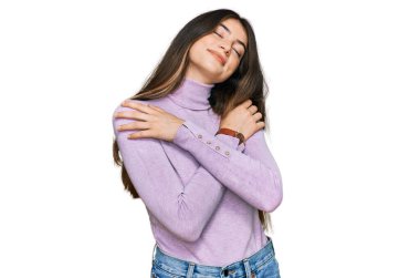 Balıkçı yaka kazak giyen genç ve güzel bir kız kendine sarılıyor mutlu ve pozitif, kendinden emin gülümsüyor. Sevgi ve özgüven. 