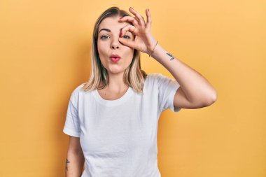 Güzel, beyaz tişört giyen beyaz bir kadın. Şaşırmış yüz ifadesiyle şok olmuş, parmaklarının arasından bakıyor. İnançsız ifade. 