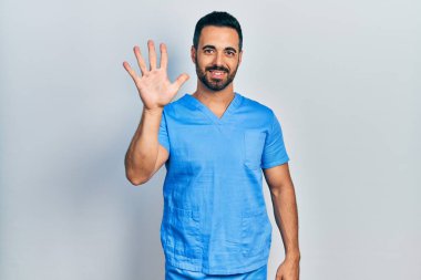 Sakallı yakışıklı, mavi erkek hemşire üniforması giyen, beş numaralı parmaklarıyla işaret eden, kendinden emin ve mutlu bir adam.. 
