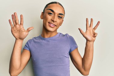 Makyaj yapan, uzun saçlı, günlük tişörtlü, dokuz numaralı parmaklarıyla gösteriş yapan, kendinden emin ve mutlu gülümseyen İspanyol adam.. 