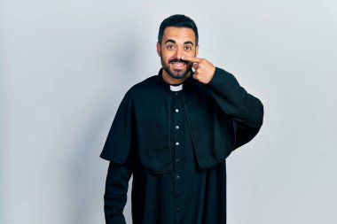 Sakallı, Katolik rahip cübbesi giyen yakışıklı bir adam. Yüzüne ve burnuna parmak işareti yapıyor. Neşeli gülümsüyor. güzellik konsepti 