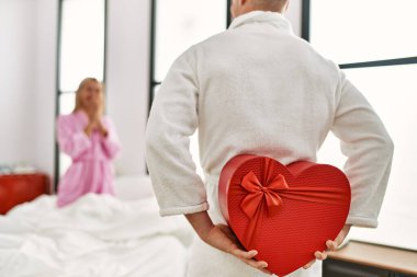 Sevgilisine sürpriz yapan bir adam sevgilisinin hediye kutusuyla evde yatağında oturuyor..