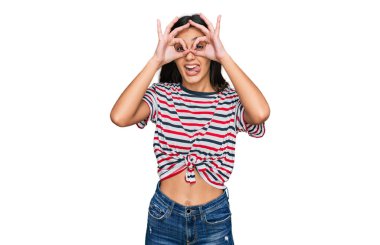 Günlük kıyafetler giyen genç İspanyol kız dürbün gibi jest yapıyor, gözleri parmaklarından bakıyor. çılgın ifade. 