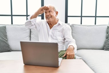 Evde dizüstü bilgisayar kullanan kıdemli bir adam koltukta mutlu bir şekilde oturuyor ve sırıtıyor. arama kavramı. 