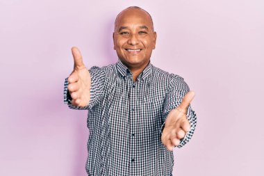 Orta yaşlı Latin bir adam, sıradan kıyafetler giyip kameraya bakıyor ve sarılmak için kollarını açmış gülümsüyor. Mutluluğu kucaklayan neşeli bir ifade.. 