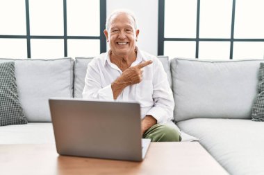 Son sınıf öğrencisi evinde dizüstü bilgisayar kullanıyor. Kanepede mutlu bir şekilde oturuyor. Yüzünde mutlu ve doğal bir ifadeyle elleri ve parmaklarıyla yüzünü gösteren bir gülümseme var. 