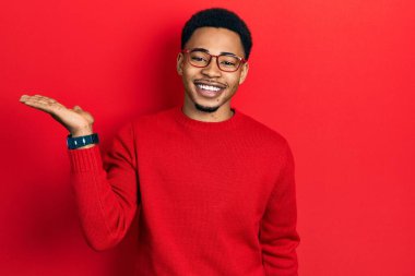 Genç Afro-Amerikan adam günlük kıyafetler ve gözlük takıyor neşeli bir şekilde gülümsüyor ve avuç içi kameraya bakıyor.. 