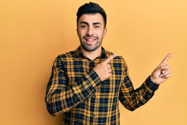 Gündelik kıyafetler giyen genç İspanyol adam gülümseyip kameraya bakıyor ve iki eliyle ve parmaklarıyla kenarı işaret ediyor.. 