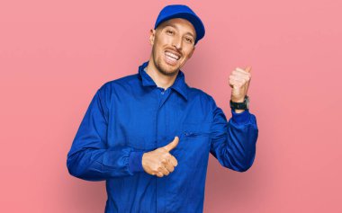 Kel, sakallı, inşaatçı tulumu giyen, arkasını gösteren, el ve baş parmakları havada, kendinden emin bir şekilde gülümseyen adam. 