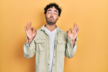 Günlük kıyafetler giyen genç İspanyol adam rahatla ve gözleri kapalı gülümse parmaklarıyla meditasyon hareketi yapıyor. yoga konsepti. 