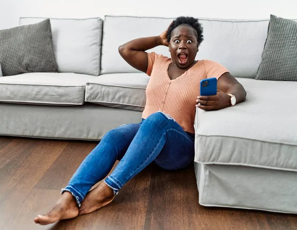 年轻的非洲女人用智能手机疯狂地坐在家里的地板上 双手放在头上吓得直哆嗦 吓得张大了嘴 吓着了一大跳 — 图库照片