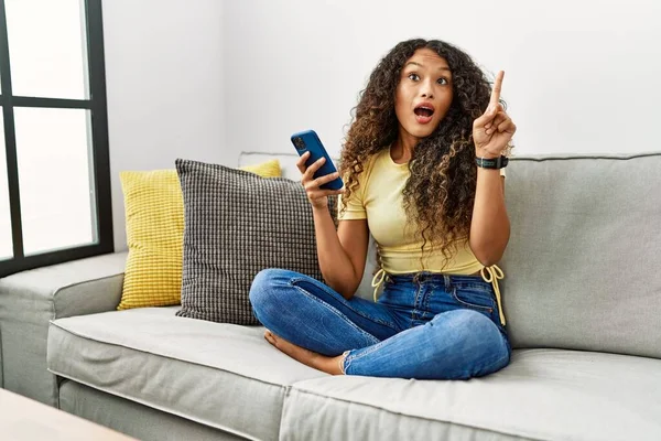 自宅のソファに座っている美しいヒスパニック系の女性は 成功したアイデアで指を指してスマートフォンを使用しています 興奮して幸せよ 第1位 — ストック写真