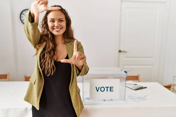 Beautiful hispanic woman standing at political campaign room smiling making frame with hands and fingers with happy face. creativity and photography concept.