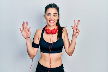 Spor kıyafetleri giyen ve kulaklık takan genç İspanyol kız sekiz numaralı parmaklarını gösterip işaret ederken kendinden emin ve mutlu bir şekilde gülümsüyor.. 