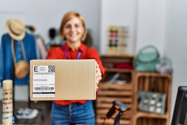 Orta yaşlı, sarışın, gülümseyen bir kadın giyim mağazasında kendine güvenen bir paket tutuyor.