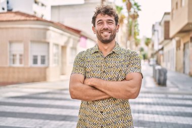 Genç İspanyol adam caddede durmuş kollarını kavuşturup kameraya bakarken gülümsüyor. pozitif kişi. 