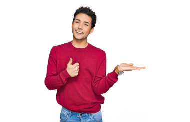 Genç ve yakışıklı bir adam günlük giysiler içinde avuçlarını gösteriyor ve baş parmaklarıyla iyi bir hareket yapıyor, mutlu ve neşeli gülümsüyor. 