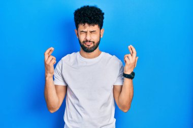 Sakallı, beyaz tişörtlü genç bir Arap. Umut dolu ve gözleri kapalı bir şekilde parmaklarıyla işaret ediyor. Şans ve batıl inançlar. 