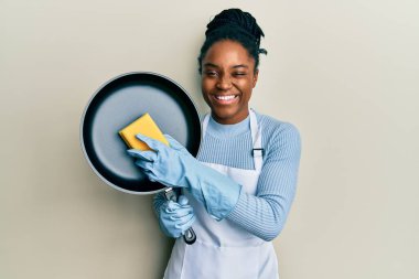 Saçları örülmüş, önlük takmış, çamaşır makinesi tutan, kameraya seksi bir ifadeyle bakan, neşeli ve mutlu yüzlü Afro-Amerikalı bir kadın.. 