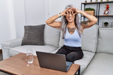 Orta yaşlı, kır saçlı bir kadın evde dizüstü bilgisayar kullanıyor. Dürbün gibi jest yapıyor. Dilini çıkarmış, gözleri parmaklarından bakıyor. çılgın ifade. 