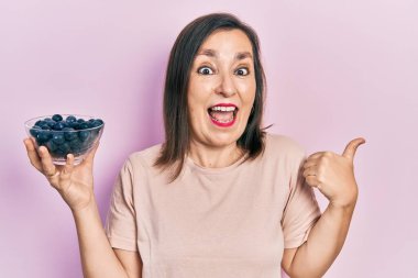 Orta yaşlı İspanyol kadın elinde yaban mersini tutarken başparmağını yana kaldırıyor ve ağzı açık gülümsüyor. 