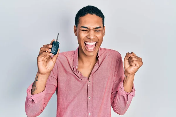 Young african american guy holding key of new car screaming proud, celebrating victory and success very excited with raised arm