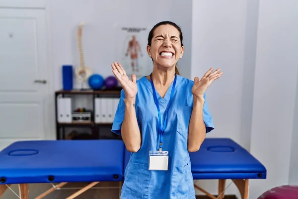 理学療法士の制服を着た若いヒスパニック系の女性が病院に立ち 腕を上げて興奮して叫んで目を閉じて成功のために狂気と狂気を祝う 勝者のコンセプト — ストック写真