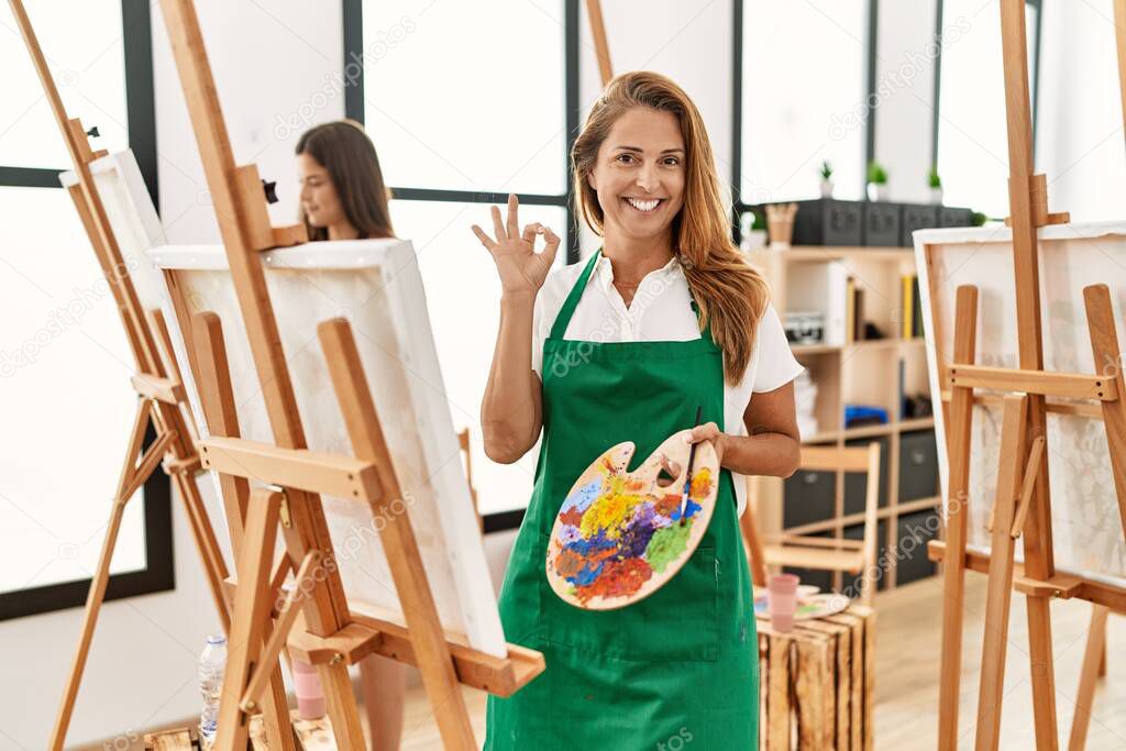 Young hispanic middle age woman at art classroom doing ok sign with fingers, smiling friendly gesturing excellent symbol 