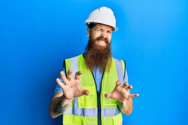 Uzun sakallı, güvenlik kaskı takan ve yansıtıcı ceketli kızıl saçlı adam tiksindirici tepki verdiği için tiksindirici ve tiksindirici bir ifade takındı. Eller havada. 